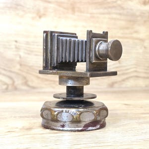 May include: A metallic sculpture shaped like a vintage bellows camera, featuring a rectangular body with a pleated accordion section and a circular lens. The object is mounted on a cylindrical base with a wide, textured, gear-like bottom rim.