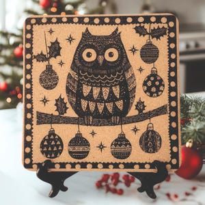 May include: A cork trivet with a black and white illustration of an owl perched on a branch with Christmas ornaments hanging from the branches. The owl has a black body with white eyes and a white beak. The ornaments are black and white with geometric patterns.