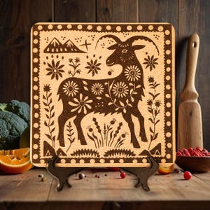 May include: A brown cork trivet with a black silhouette of a goat surrounded by flowers and mountains. The trivet has a decorative border of dots and a scalloped edge.