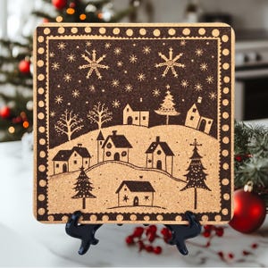 May include: A cork trivet with a black and white illustration of a winter village scene. The trivet is square and has a black border with white polka dots. The illustration features houses, trees, and snowflakes.