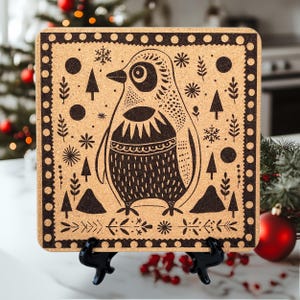 Puede incluir: Un posavasos de corcho con una ilustración en blanco y negro de un pingüino rodeado de una escena de bosque invernal. El posavasos tiene un fondo de corcho marrón y un borde negro con lunares blancos.