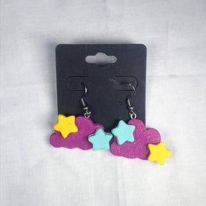 May include: A pair of handmade earrings featuring a cloud and star design. The earrings have a purple glitter cloud shape with yellow and light blue stars. The earrings are attached to silver-colored fish hook ear wires.