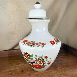 May include: White ceramic vase with a lid, decorated with red, green, and blue floral designs. The vase has a gold trim around the lid and base.