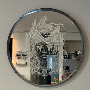 Może przedstawiać: Okrągłe lustro w srebrnej ramie z grawerowanym wzorem twarzy kobiety ze wężami we włosach. Kobieta ma łzy spływające po policzkach.