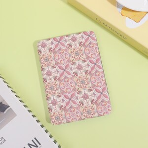 May include: A rectangular notebook featuring a floral design in pink, red, and beige. The notebook is placed on a light green surface, with a book and a box with the text "LA PATISSERIE" in the background.