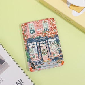 May include: A rectangular notebook with a colorful illustration of a bookstore facade. The image features a brick building with windows, a bicycle, and the text "THE BOOKSHOP". The design includes floral accents and a welcoming outdoor seating area.