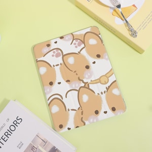 May include: A white tablet case with a repeating pattern of cartoon corgi faces in shades of tan and brown. The case has a gold-colored clasp. The background is a light green color.
