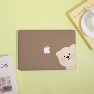 May include: A brown laptop with a white Apple logo and a cartoon bear peeking from the side. The bear has a blush pink cheek and black eyes. The laptop is on a light green surface.