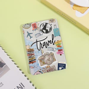 May include: A travel-themed notebook featuring a world map design, decorated with stickers of a globe, airplane, suitcase, and camera. The word "Travel" is written in a cursive font. Includes city name stickers and a passport.