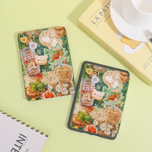 May include: Two tablet cases with a whimsical design featuring cartoon bears, bunnies, and food items like strawberries and cookies. The cases have a green background with a variety of stickers and illustrations. One case is slightly angled, showing the design more clearly.