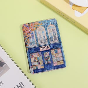 May include: A rectangular, blue book cover featuring a detailed illustration of a bookstore. The illustration includes a storefront with the text "BOOK SHOP" above the entrance, and a tree with orange and pink leaves. The windows are white.