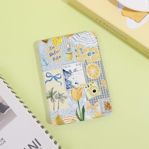 May include: A rectangular notebook with a collage design featuring lemons, a camera, and the text "La Dolce Vita." The notebook has a blue and yellow colour scheme with images of Italy and palm trees.