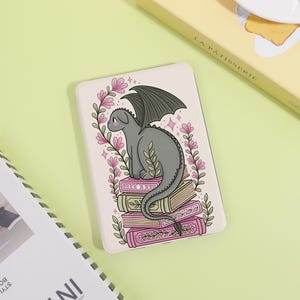 May include: A rectangular e-reader case featuring a gray dragon perched atop a stack of four books. The illustration includes pink floral vines framing the dragon and books, with small pink star accents on a light cream-colored background.