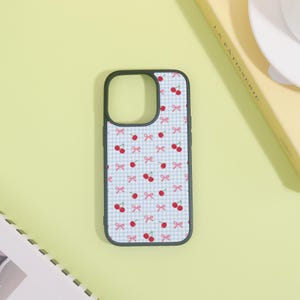 May include: A black phone case with a light blue gingham pattern featuring red cherries and pink bows. The case has a cutout for the camera and buttons. The design is repeated across the entire surface of the case.