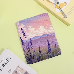 May include: A tablet case with a landscape design featuring lavender flowers, mountains, and a sunset sky in shades of purple, pink, and blue. The case is rectangular with rounded corners. A book and a dessert box are also in the image.