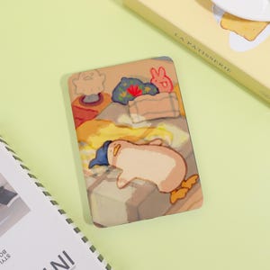 May include: A rectangular, illustrated phone case featuring a sleeping penguin wearing a blue hat. The case has a beige background with a bed design, including a pillow and a rabbit. The case is next to a book with the words "LA PATISSERIE".