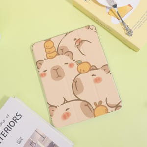 May include: Beige tablet case featuring a repeating pattern of cartoon bear faces with blush cheeks and fruit accents. The case is open, revealing the screen. The background is a light green.