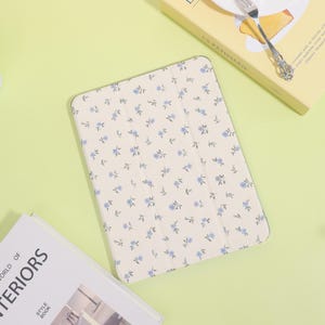 Peut inclure: Étui de tablette crème avec un motif floral de petites fleurs bleues et de feuilles vertes. L'étui est ouvert. Un livre et une boîte jaune avec du texte sont en arrière-plan.