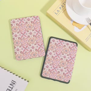 K&ouml;nnte beinhalten: Zwei Tablet-H&uuml;llen mit einem dekorativen Blumenmuster in Rosa-, Rot- und Cremet&ouml;nen. Die H&uuml;llen haben einen schwarzen Rand und werden auf einer hellgr&uuml;nen Oberfl&auml;che pr&auml;sentiert. Eine Schachtel mit dem Text "LA P&Acirc;TISSERIE" befindet sich im Hintergrund.