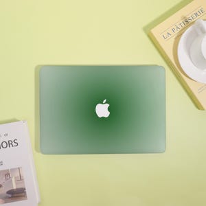 May include: A green laptop with a white Apple logo in the center. The laptop is on a light yellow surface, with a book and a book with a cup and saucer visible in the background.