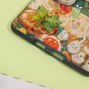 May include: A phone case with a colorful illustration of food items, including bread, strawberries, and a pastry. The case has a dark gray border and a detailed, cartoon-like design with a light green background.