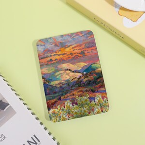May include: Rectangular art print featuring a colorful landscape painting of rolling hills and a vibrant sunset. The artwork showcases a blend of warm and cool tones, with a focus on the natural scenery. The print is on a light green surface.