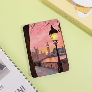 May include: A rectangular art print featuring a London cityscape at sunset. The image showcases the Houses of Parliament, Big Ben, and a lamppost. The sky is a blend of pink and orange hues, with a river in the foreground.