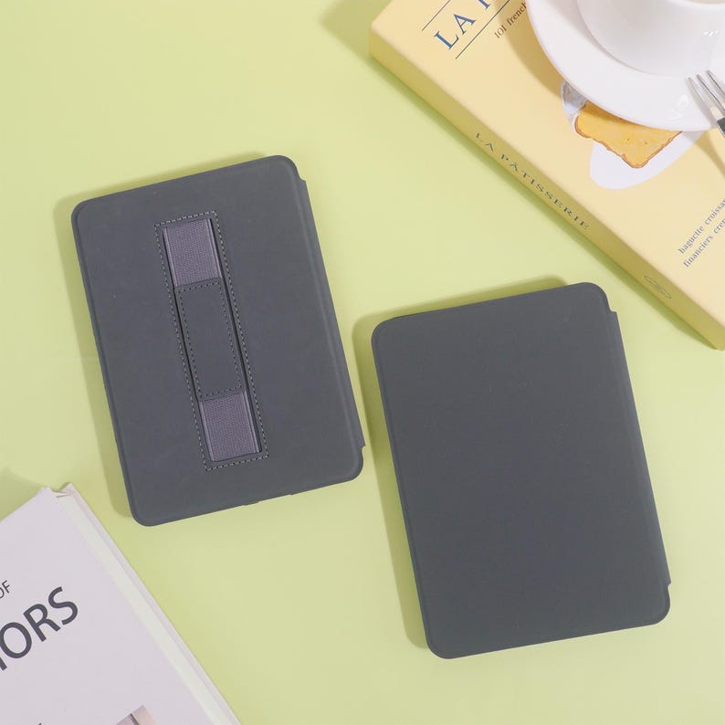 May include: Two gray tablet cases are displayed on a light green surface. One case has a gray elastic strap for holding. A book and a box with the text "LA PATISSERIE" are also visible.