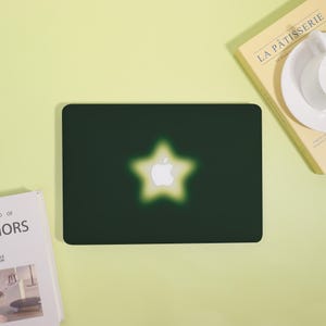 May include: Dark green laptop case featuring a glowing star design with the Apple logo at the center. The star has a soft, yellow-green gradient. The case is rectangular and sits on a light green surface.