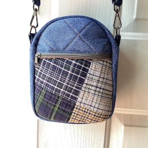 May include: A crossbody bag featuring a quilted blue denim upper section and a lower front panel made of patchwork plaid fabric in shades of navy, green, white, and tan. The bag includes a horizontal zippered pocket and silver metal hardware.