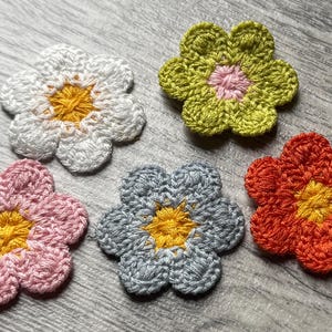Może przedstawiać: Pięć ręcznie robionych aplikacji kwiatowych na szydełku w różnych kolorach: białym, różowym, zielonym, szarym i pomarańczowym. Każdy kwiat ma żółty środek. Te dekoracyjne ozdoby kwiatowe są idealne do projektów rzemieślniczych.
