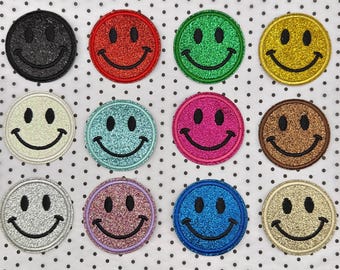 Könnte beinhalten: Eine Sammlung von zwölf glitzernden Smiley-Aufnähern in verschiedenen Farben, darunter Schwarz, Rot, Grün und Gold. Jeder Aufnäher hat ein schwarz gesticktes Lächeln und Augen, ideal zum Aufpeppen von Kleidung oder Accessoires.