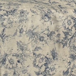 Op de afbeelding: Crèmekleurige stof met een herhalend patroon van blauwe bloemmotieven. Het patroon bevat gedetailleerde illustraties van bloemen en bladeren, wat een vintage esthetiek creëert. De stof lijkt een katoen- of linnenmix te zijn.