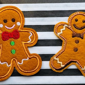 May include: Two gingerbread man felt patches. The left patch features a red bow tie, white icing details, and two green buttons. The right patch has a brown bow tie, white icing accents, and two brown buttons. Both have embroidered facial features.