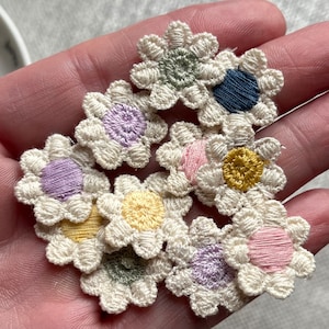 May include: A collection of small, embroidered daisy flower patches. Each flower features a cream-colored petal design with a colorful center in shades of purple, yellow, pink, blue, and green. These patches are ideal for adding a decorative touch to clothing or crafts.