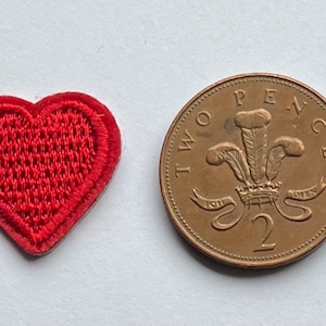 May include: A red heart-shaped embroidered patch, next to a two pence coin. The patch has a detailed outline and a woven appearance. The coin is bronze-coloured and features a design with text and a crown.
