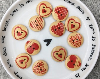 Op de afbeelding: Houten knopen met rode hartpatronen op een wit bord. De knopen tonen diverse hartontwerpen, waaronder strepen en stippen. Het bord heeft tekst rond de rand, waaronder de woorden "love is kind."