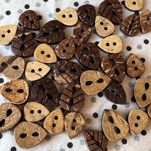 Può includere: Una collezione di bottoni in legno naturale di varie forme a foglia e a goccia. I bottoni sono in tonalità marroni e beige, con due o quattro fori per cucire. I bottoni sono sparsi su una superficie bianca con pois neri e grigi.