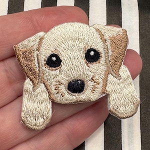 Pode incluir: Patch bordado com um cachorrinho bege claro com orelhas e detalhes castanhos. O cachorrinho tem olhos grandes pretos e um nariz preto. O patch foi projetado para ser costurado ou passado a ferro no tecido, ideal para personalizar roupas ou acessórios.