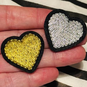 May include: Two heart-shaped patches. One is gold with a black border, the other is silver with a black border. The patches are on a striped background. These patches could be used for clothing or accessories.