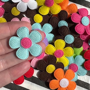 May include: A collection of colorful felt flower appliques in various shades including blue, pink, yellow, orange, brown, and white. Each flower has a contrasting center and is arranged on a striped background.