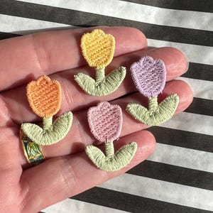 May include: Four embroidered tulip patches in orange, yellow, pink, and purple. Each patch features a detailed flower head, green stem, and leaves. The patches are held in a hand against a black and white striped background.