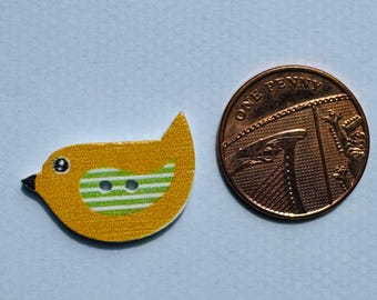 May include: A yellow bird-shaped button with green and white stripes, black eye and beak, and two holes for sewing. Next to it is a copper One Penny coin.
