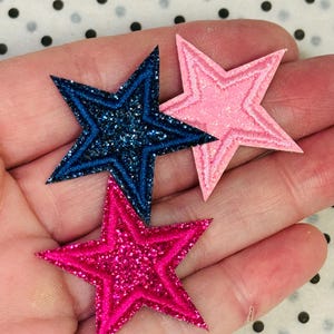 May include: Three star-shaped patches in shades of pink and blue. The blue star has glitter, the pink stars have embroidered edges. The patches are on a dotted background.