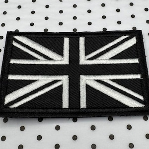 May include: A rectangular black and white embroidered patch featuring the Union Jack flag design. The flag's cross and diagonal stripes are white against a black background. The patch is likely for clothing or accessories.