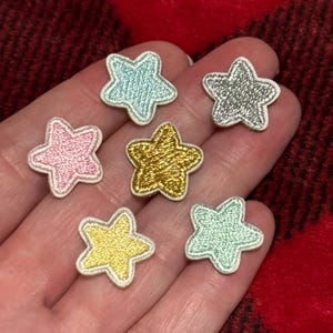 Può includere: Sette toppe ricamate a forma di stella nei colori rosa, oro, argento e blu. Ogni stella ha un bordo bianco. Le toppe sono presentate su uno sfondo a quadri rossi e neri.