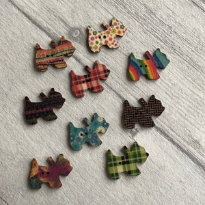 Könnte beinhalten: Eine Sammlung von neun holzgeschnitzten Knöpfen in Hundeform mit verschiedenen Mustern. Die Muster umfassen Blumen-, Karo- und geometrische Designs in verschiedenen Farben. Jeder Knopf hat zwei kleine Löcher zum Annähen.