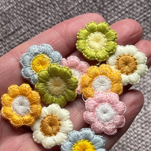 May include: A collection of small, embroidered flower appliques in various pastel colors. The flowers have yellow, white, pink, and green petals with yellow or brown centers. The appliques are likely for crafting or embellishment.