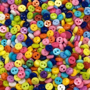 Könnte beinhalten: Eine lebendige Auswahl an kleinen, runden Knöpfen in verschiedenen Farben, darunter Rosa, Gelb, Blau, Grün und Orange. Jeder Knopf hat zwei oder vier Löcher zum Annähen. Die Knöpfe sind verstreut und bilden ein farbenfrohes und strukturiertes Muster.