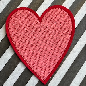 May include: A red heart-shaped patch with a red border. The heart has a textured appearance and is set against a black and white striped background.